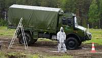 Ein Soldat in ABC-Schutzausstattung steht mit einem Sprühschlauch vor einem UNIMOG-Lkw