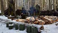 Im Vordergrund liegen gestapelte Holzscheite für ein Lagerfeuer, dahinter bauen Soldaten das Biwak.