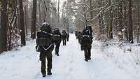 Soldaten mit Rucksäcken marschieren in zwei Reihen versetzt auf einem verschneiten Weg im Wald.