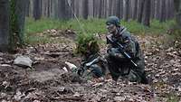 Ein Soldat steht getarnt in einer tiefen Stellung im Wald, ein anderer Soldat hockt neben ihm.