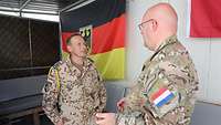 Im Hintergrund befindet sich eine Sitzecke und eine Deutschlandflagge. Im Vordergrund sprechen zwei Soldaten 