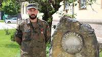 Ein Soldat steht neben einem Bataillonsstein in der Sonne und schaut in die Kamera.
