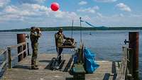 Zwei Soldaten, daneben ein roter Ballon stehen auf dem Steg am Schwielowsee. Einer funkt, der zweite beobachtet die Landezone