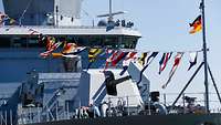 Bunte Flaggen wehen über das Deck eines grauen Schiffes.