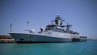 A grey warship berthed at a harbour’s pier.