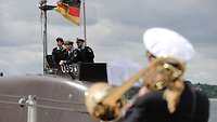 Der dunkelgraue Turm eines U-Boots mit drei Marinesoldaten in blauer Arbeitsuniform; davor eine Musikerin mit Posaune.
