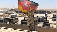 Eine Soldatin steht neben der Landesflagge von Niedersachsen. Im Hintergrund Stromerzeugungscontainer 