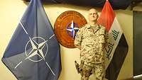 Ein deutscher Soldat steht vor zwei Flaggen. Im Hintergrund befindet sich eine Holzschnitzerei