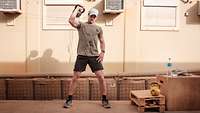 Ein Soldat steht in Sportzeug vor einem Gebäude und stemmt einen Kettlebell aus Eisen