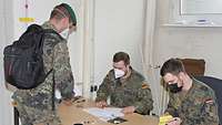 An einem Tisch sitzen zwei Soldaten und überprüfen die Unterlagen eines dritten Soldaten. Dieser steht vor dem Tisch.