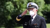 Ein Marineoffizier in dunkelblauer Uniform und weißer Schirmmütze hat die Hand zum militärischen Gruß erhoben.