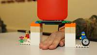 Unter einer Legobrücke liegt eine Hand auf dem Tisch und auf der Brücke steht eine Flasche.
