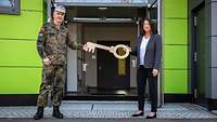 Vor einem gelbgrünen Eingangsbereich überreicht eine Frau einem Soldaten symbolisch den Schlüssel.