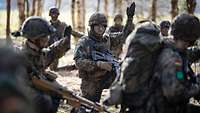 Mehrere Soldaten hocken im Wald zusammen. Eine Soldat in der Mitte hebt dabei die Hand.