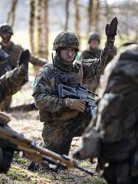 Mehrere Soldaten hocken im Wald zusammen. Eine Soldat in der Mitte hebt dabei die Hand.