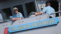 Zwei Marinesoldaten in blauer Arbeitsuniform in der Nock eines grauen Schiffes; einer von beiden trägt eine weiße Schirmmütze.