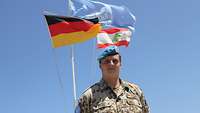 Ein Soldat steht vor der VN-Flagge, der libanesischen und der deutschen Flagge