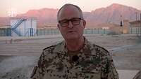 Generalinspekteur Zorn im Porträt spricht in die Kamera, im Hintergund ein Feldlager der Bundeswehr vor einem Gebirgszug