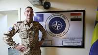 Ein Soldat steht vor einer Wand. Hinter ihm eine Tafel mit dem Wappen der NATO Mission Iraq