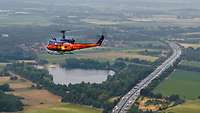 Ein bunt lackierter Hubschrauber überfliegt eine vierspurige Autobahn, auf der Fahrzeuge fahren.