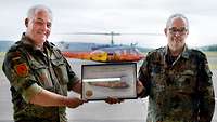 Zwei Soldaten halten zwischen sich ein gerahmtes Foto, im Hintergrund steht ein rot-gelb lackierter Hubschrauber.