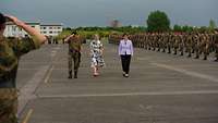 Zwei Frauen und ein militärisch grüßender Soldat laufen an vielen Soldatinnen und Soldaten vorbei