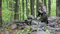 Ein Soldat liegt mit seiner Waffe im Anschlag in Stellung. Neben ihm hockt ein Ausbilder.