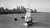 Auf einem schwarz-weiß-Bild segelt ein Schiff vor der Sillouette von New York über das Wasser.