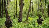 Drei Soldaten knien in einem Waldstück. Zwei von ihnen sichern die Umgebung. Ein dritter befestigt einen Lautsprecher