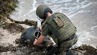 A service member in battle dress wearing a helmet kneels on the beach, defusing a sea mine.