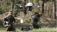 An airborne UAV in the foreground and two service members, one of them controlling the UAV.