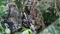 Two service members in camouflage uniform hiding in a wooded area, carrying rifles in their hands.