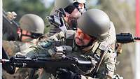 A service member in battle dress peeking around the corner of a house, holding an assault rifle in firing position.