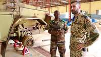 Ein deutscher und ein malischer Soldat stehen nebeneinander im Gespräch. Neben ihnen steht ein Militärfahrzeug zur Reparatur