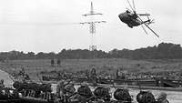 Schwarz-weiß-Foto: Hubschrauber fliegt schräg durch die Luft. Am Boden sind Soldaten zu sehen.