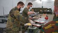 Zwei Soldaten stehen an einer Verpflegungsstraße und geben das Essen aus Thermen an Soldaten aus