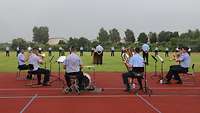 Mehrere Personen sitzen zusammen und spielen Musikinstrumente. Im Hintergrund sind die angetretenen Soldaten zu sehen.