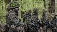 Soldaten mit Helm, Tarnschminke im Gesicht und Waffe stehen im Wald in einer Reihe und werden unterrichtet