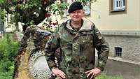 Neben einem Gebäude steht ein Stein mit einem Wappen, daneben ein Soldat in Uniform.