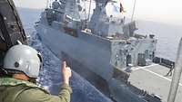 Ein Soldat in einem Hubschrauber zeigt einen Daumen hoch in Richtung der Besatzung eines Schiffes