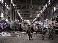 Drei Soldaten in ABC-Schutzkleidung in der Halle einer Industrieruine vor vier Tankbehältern mit Graffiti besprüht 