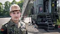 Soldat mit Helm steht vor einer abgetarnten Richtfunkkabine, seinem Arbeitsplatz als Netzwerkadministrator.