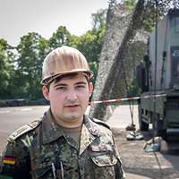 Soldat mit Helm steht vor einer abgetarnten Richtfunkkabine, seinem Arbeitsplatz als Netzwerkadministrator.