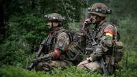 Zwei Soldaten hocken im Wald, der rechte spricht in ein Funkgerät, der linke sichert ihn mit einem Gewehr.