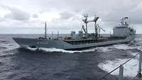 A long and slender grey ship is underway in rough seas.
