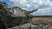 Ein Panzer mit einem Tarnnetz bespannt steht auf sandigem Boden. Das Rohr zeigt in Richtung einer bewaldeten Fläche