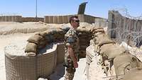 Ein Soldat steht vor einem Hesco-Schutzwall und schaut in die Ferne. . Vor ihm liegen mehrere Sandsäcke 