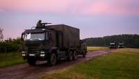 Militärische Lkw-Kolonne auf einem Truppenübungsplatz in der Abenddämmerung. Aus dem Dach lugt jeweils ein Soldat mit Gewehr.