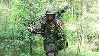 Ein Scharfschütze geht aufrecht durch einen Mischwald. Sein Gewehr trägt er über der Schulter.