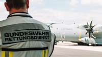 Ein Angehöriger des Rettungsdienstes steht im Vordergrund mit dem Rücken zum Fotografen. Im Hintergrund steht ein Flugzeug.
